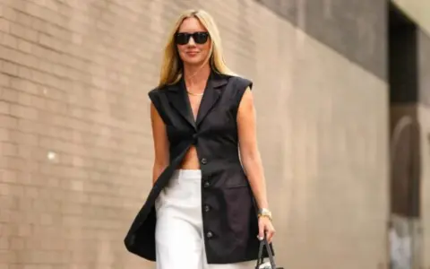 Donna con un elegante outfit con bermuda bianchi e blazer nero senza maniche, occhiali da sole e borsa.
