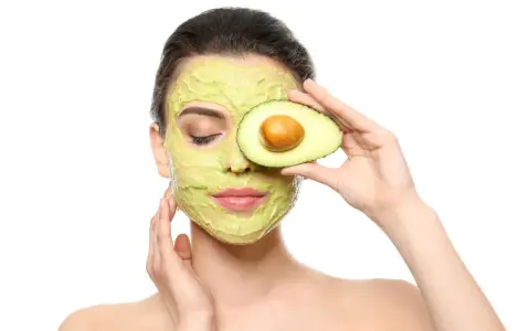 Frau mit Avocado-Gesichtsmaske und Avocado-Scheibe vor dem Auge. Die Avocado-Wirkung auf die Haut ist bekannt für ihre pflegenden Eigenschaften.