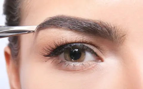 Une pince à épiler retire délicatement un poil du sourcil, affinant la ligne pour un regard parfait.