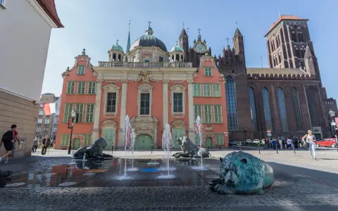 Fontanna Czterech Kwartałów w Gdańsku z lwami i wodnymi strumieniami przed różową kamienicą i bazyliką.
