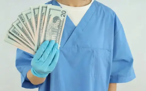 Higienistka stomatologiczna w niebieskim fartuchu pokazuje wachlarz banknotów 20 dolarowych. Zarobki netto mogą być satysfakcjonujące.