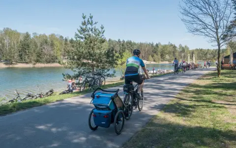 Rodzina na rowerach z przyczepką nad jeziorem Świerklaniec. W tle inni rowerzyści i plażowicze.
