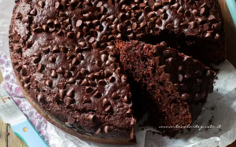 Torta al cioccolato senza uova, soffice e golosa, ricoperta di gocce di cioccolato. Una fetta mostra la sua morbidezza.