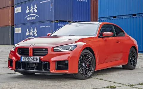Pomarańczowe BMW M2 z czarnymi felgami, na tle niebieskich kontenerów. Sprawdź, jaka jest aktualna bmw m2 cena.