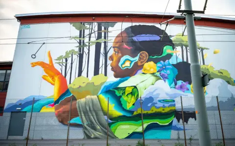 Färgstark street art på en vägg visar en person i profil med en fjäder i håret, omgiven av träd och blommor.