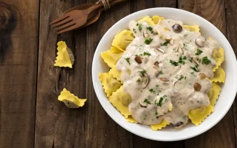 Ravioli w kremowym sosie śmietanowym z grzybami i natką pietruszki, podane na drewnianym stole.