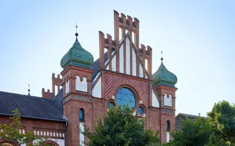 Das imposante Gebäude im Stil des Zollern-Schlosses mit seinen grünen Kuppeln und roten Ziegeln ragt vor blauem Himmel auf.