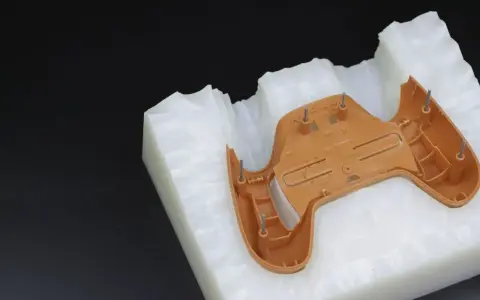 A brown plastic part, likely from a game controller, is placed on a white, textured material. This setup suggests a step in the polyurethane vacuum casting process, where a mold is being prepared.