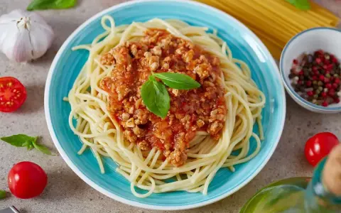 Pyszne spaghetti z bogatym sosem bolognese, udekorowane listkiem bazylii. Obok czosnek, pomidorki i przyprawy.