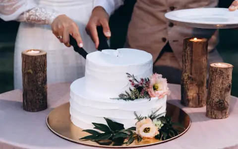 Biały tort weselny z teksturą, ozdobiony kwiatami. Para kroi tort, a obok płoną świece w drewnianych świecznikach. Trendy w prezentacji tortów weselnych to m.in. tekstury.