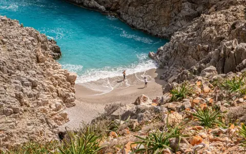 Malownicza zatoka z turkusową wodą i piaszczystą plażą, otoczona skalistymi klifami. Dwie osoby spacerują po brzegu. Chania plaże zachwycają.