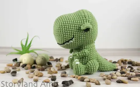 A green crocheted dinosaur sits on a white surface surrounded by small stones and a small plant.