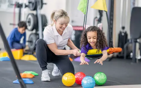 Ergotherapie für Kinder: Ein Mädchen lacht auf einer Schaukel, während eine Therapeutin sie unterstützt. Bunte Bälle liegen im Vordergrund.
