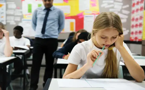 Uczennica zamyślona nad arkuszem, zastanawia się, ile trzeba mieć procent żeby zdać maturę. Nauczyciel obserwuje klasę.