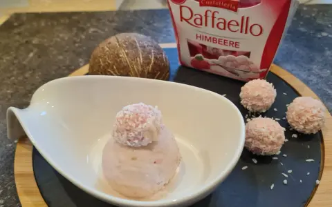 Ein Löffel Raffaello Himbeer Eis in einer Schale, daneben drei Raffaello-Kugeln und eine Kokosnuss.