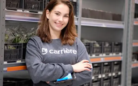 Young woman in a grey sweatshirt with "carrière" logo, holding a scanner, in a warehouse with shelves of black crates.