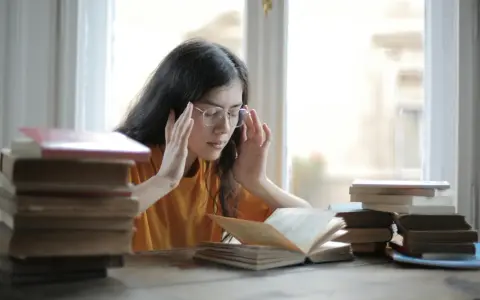 Ragazza con occhiali e mani sulle tempie, circondata da libri. Potrebbe soffrire di mancanza di concentrazione cause.