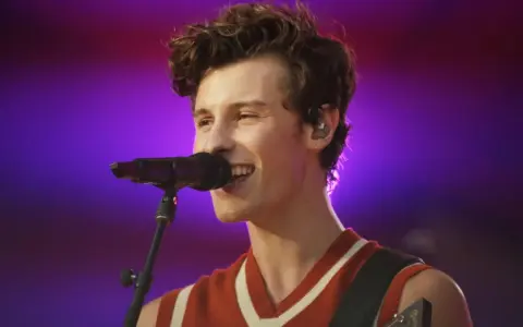 Shawn Mendes śpiewa swoje utwory na koncercie, z mikrofonem przed ustami i gitarą na ramieniu.