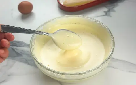 Préparation d'un tiramisu : une cuillère de pâte à bombe aérienne est prélevée d'un bol en verre. Un œuf et des biscuits à la cuillère sont visibles.