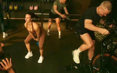 Träningspass med fokus på kondition och styrka. En kvinna gör knäböj med kettlebell, en man kastar medicinboll och en annan cyklar på en Air Bike. Kanske en övning för att hålla gunde svan vilopuls nere.