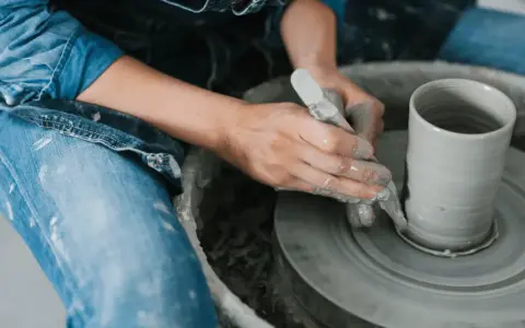 Händer skulptera i lera på en drejskiva. En person i jeans och jeansjacka formar en cylinder.
