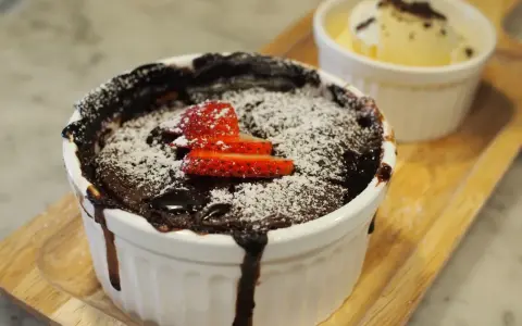 Pyszny, gorący lava cake z mikrofali, posypany cukrem pudrem i udekorowany truskawkami. Obok porcja lodów waniliowych.