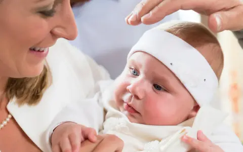 Uśmiechnięta mama trzyma dziecko w białej szatce. Dłoń ojca błogosławi malucha. Zastanawiasz się, ile kosztuje chrzest?
