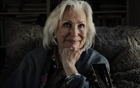 Äldre kvinna med grått hår, gitarr och en fluffig pläd. Trots att Marie Bergman är sjuk, lyser hennes ögon av liv.