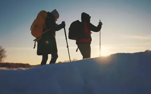 Dwie osoby z plecakami i kijkami trekkingowymi idą przez ośnieżony teren. W tle zachodzące słońce, przypominające czerwone wierchy szczyty.