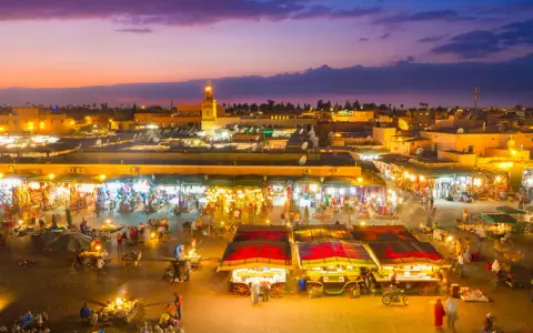 Der belebte marrakesch marktplatz erwacht bei Dämmerung zum Leben. Stände mit bunten Waren, Essensverkäufer und Menschenmassen beleben die Szene unter dem lila Himmel.