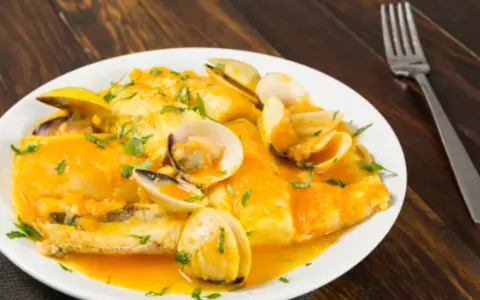 Merluza a la marinera con almejas, servida en un plato blanco sobre una mesa de madera oscura.