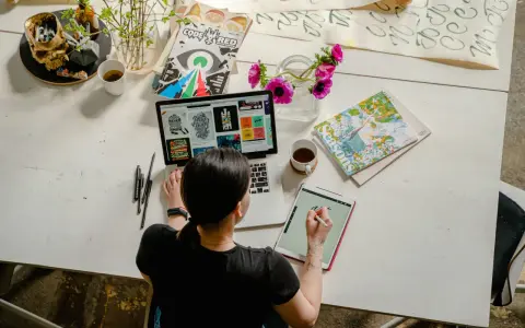 Kobieta pracuje online w domu, projektując na laptopie i tablecie. Na biurku kwiaty, kubek kawy i czaszki z pisakami.