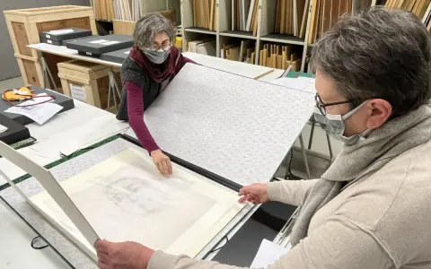 Deux personnes manipulent un dessin au crayon, peut-être un portrait, sur une table. Le vernis protège l'œuvre.