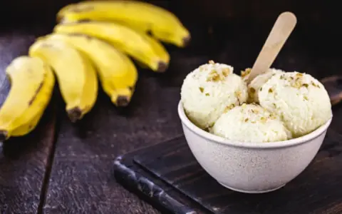 Delicioso helado de plátano cremoso, adornado con nueces, junto a racimos de plátanos frescos sobre una mesa de madera rústica.