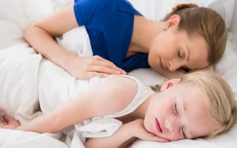 Mama i dziecko śpią razem, otuleni białymi pościelami. Czy dzieci powinny spać z rodzicami? Ta chwila spokoju sugeruje, że tak.