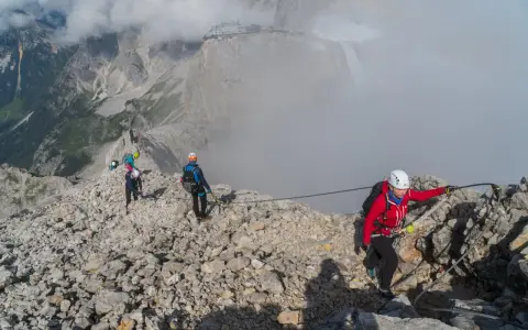 Grupa turystów pokonuje via ferrata polska. Wspinają się po skalistym grzbiecie, asekurowani linami. W tle widać góry i budynek na szczycie.
