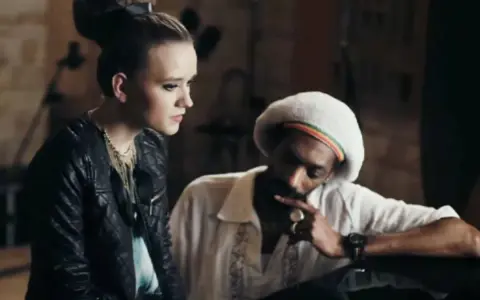 Snoop Dogg w białej koszuli i beretce Rasta, rozmawiający z młodą kobietą. Może to być scena z jednego z jego **snoop dogg utworów**.