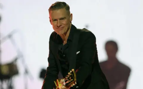 Bryan Adams w czarnym garniturze gra na gitarze. Sprawdź jego nadchodzące wydarzenia i koncerty.