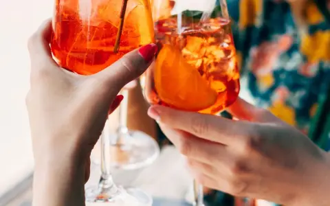 Zwei Hände halten Aperol Spritz Gläser mit Eis und Orangenscheiben. Ein perfektes Aperol Rezept für den Sommer.