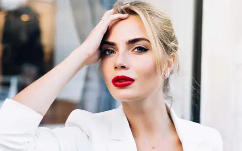 Frau mit roten Lippen und weißem Blazer, Hand am Kopf.