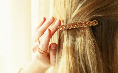 Złota klamra w kształcie łańcuszka ozdabia blond włosy. Dłoń z pierścionkami delikatnie je układa, pokazując, jak upiąć włosy klamrą.