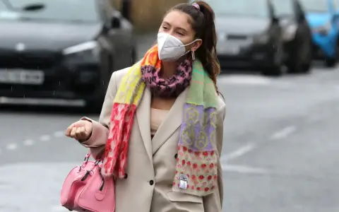 Iris Law, con mascarilla y bufanda multicolor, camina por la calle con un bolso rosa.