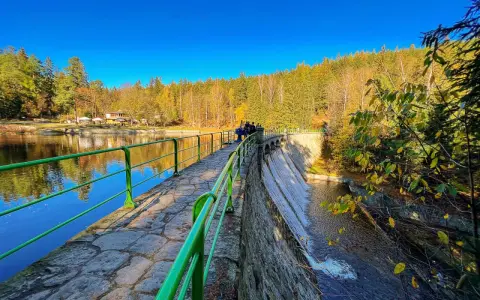 Kamienna zapora z zieloną balustradą, wodospadem i jesiennym lasem w tle.