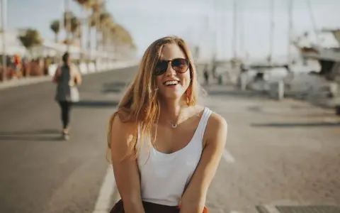 Glückliche junge Frau mit Sonnenbrille und weißem Top genießt ihr Praktikum in Spanien am Hafen.