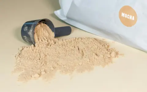 Moccha-smakande proteinpulver spillt från en skopa, med påsen i bakgrunden. Perfekt för att hålla koll på proteinpulver kcal.
