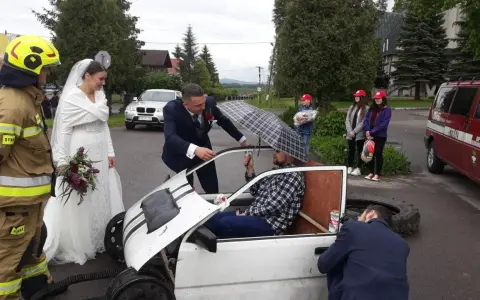 Brama weselna z udziałem strażaka, pana młodego i panny młodej. Pan młody rozmawia z druhem siedzącym w prowizorycznym samochodzie.