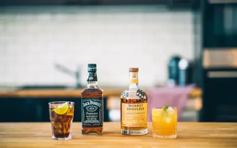 Zwei Flaschen Whiskey, Jack Daniel's und Monkey Shoulder, stehen neben zwei Cocktails auf einem Holztisch.