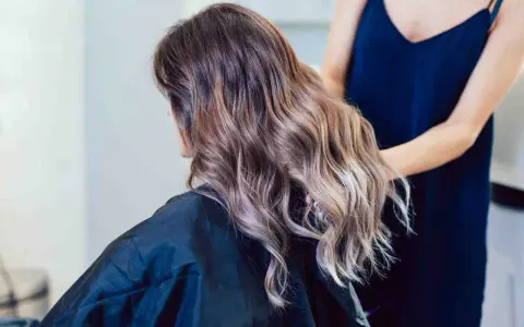 Kobieta z falowanymi włosami w odcieniach brązu i subtelnym balejażem blond. Fryzjerka poprawia jej fryzurę.