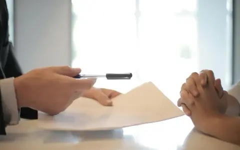 Dwie osoby przy stole, jedna podaje dokumenty, druga z dłońmi złożonymi w geście oczekiwania. Czy umowa zlecenie wlicza się do emerytury?