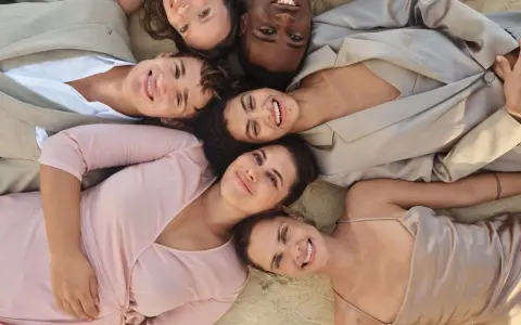 Sechs lächelnde Menschen liegen eng beieinander. Ihre Kleidung, wie ein beiger Anzug und ein rosafarbenes Kleid, inspiriert die Idee einer leichten basisgarderobe sommertyp.